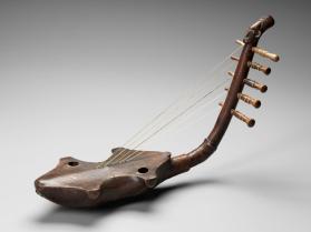 Medium-brown arched harp with two holes in base and strings that connect up the arch to five tuning pegs. Base is an anthropomorphic head