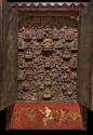 Red wooden cabinet with doors open showing many, closely-packed ceramic votive figures inside. Small depiction of people painted at the bottom
