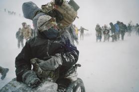 Photo of figures bundled in cold-weather gear during a blizzard; one kneels in the foreground wearing a large backpack