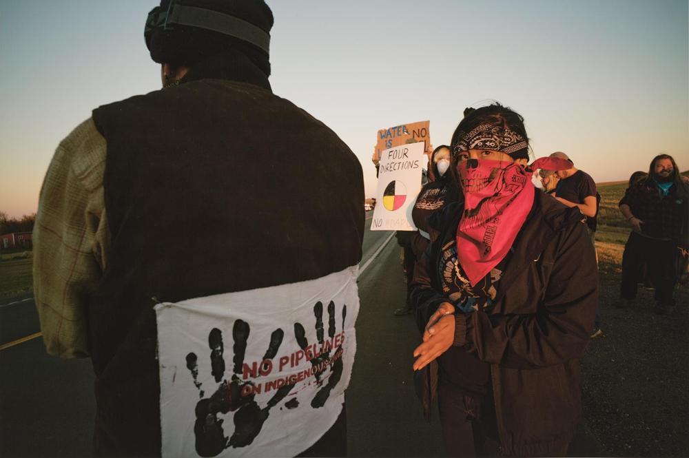 Photo of people gathered by a road. One figure with a sign on their back says “no pipeline on indigenous land.” Another has a red bandana face mask