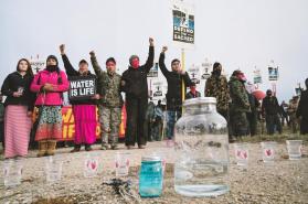 Photo of people with raised fists holding signs that say “water is life.” Figures stand behind water vessels of various sizes
