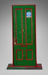 Bullet-pocked green door. Trim painted in red. A small blue half circle on the upper right contains 4 white stars and words, “U.S Approved”